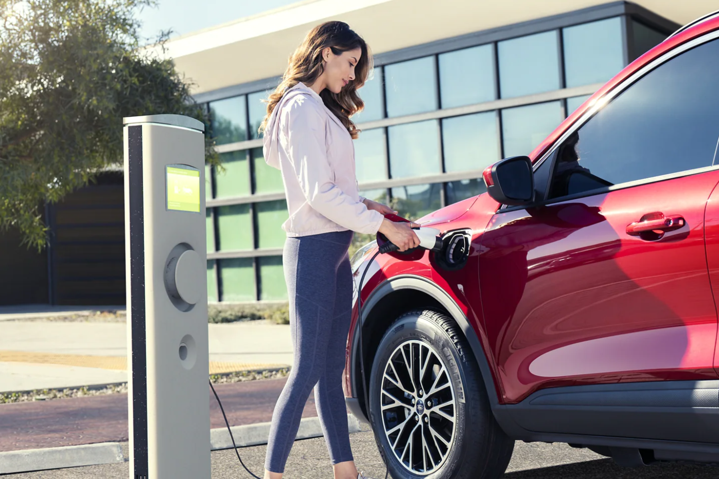 A woman plugs in her hybrid, red 2022 Ford Escape to a charging station in an urban setting on a sunny day.