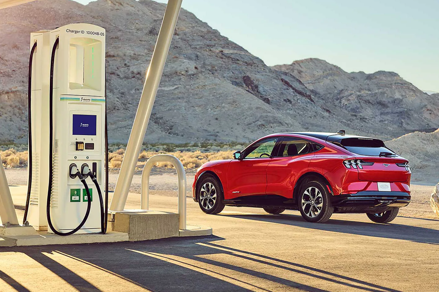 A red 2022 Ford Mach-E sit parked facing away from the viewer at a charging stations as the sun sets and casts long shadows in a desert scene