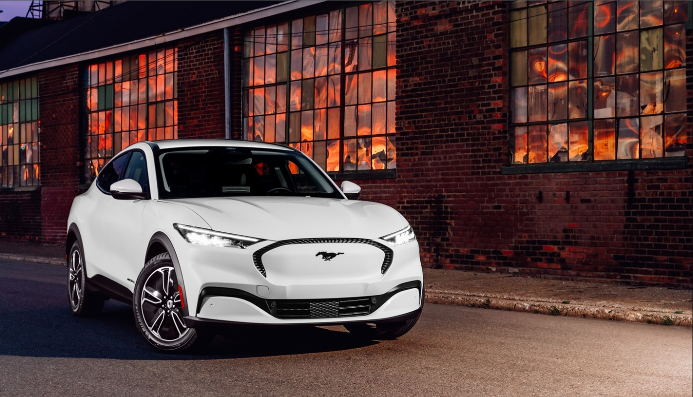 A front, angle view of a white 2022 Ford Mach-E showing its mostly solid grille as it sits parked in front of a brick building