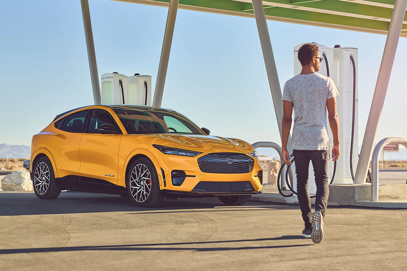 A man walks towards a yellow 2022 Ford Mustang Mach-E parked at an EV charging station as the sun sets