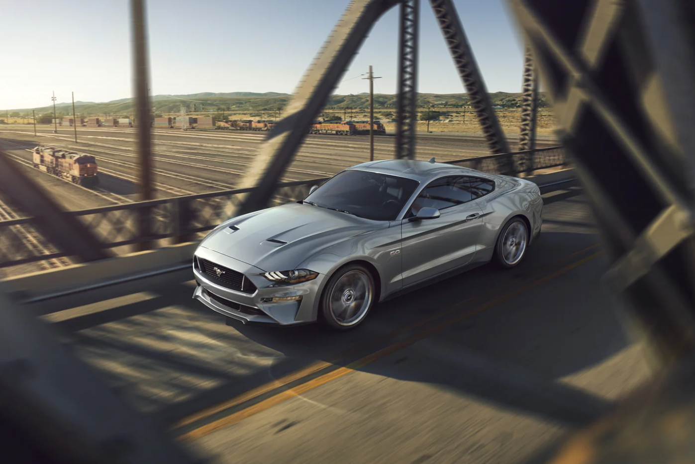 A silver 2022 Ford Mustang speeds past a train depot on a bridge